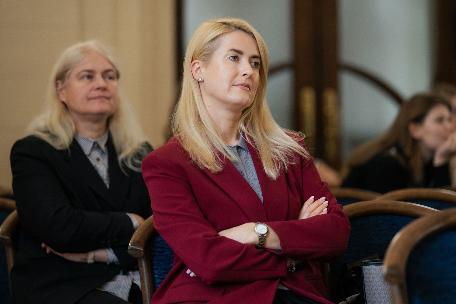 Two women sit in the conference audience, one in a burgundy blazer with crossed arms and one in a black jacket.