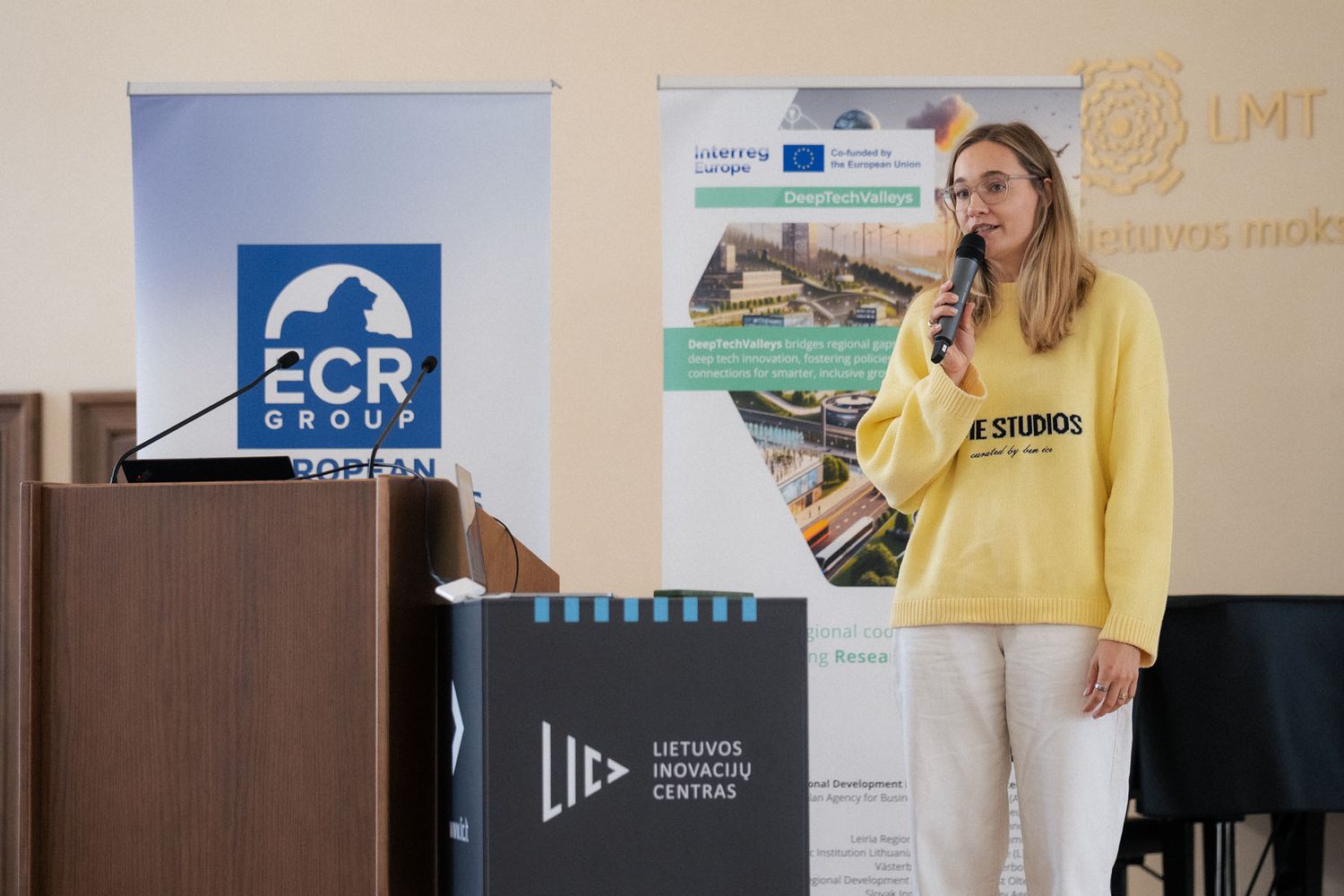 Woman in a yellow sweater speaks into a microphone on stage beside ECR Group banners and a Lithuanian Innovation Centre box.