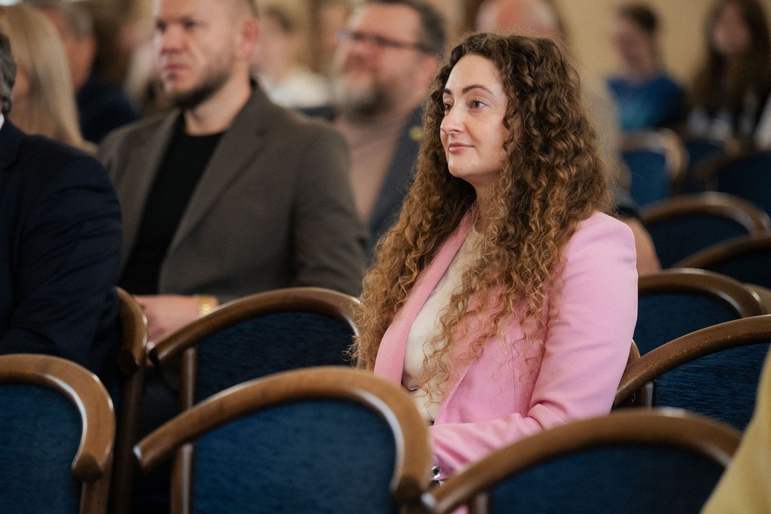 A woman with long curly hair and a pink blazer sits attentively in a blue chair among conference attendees.