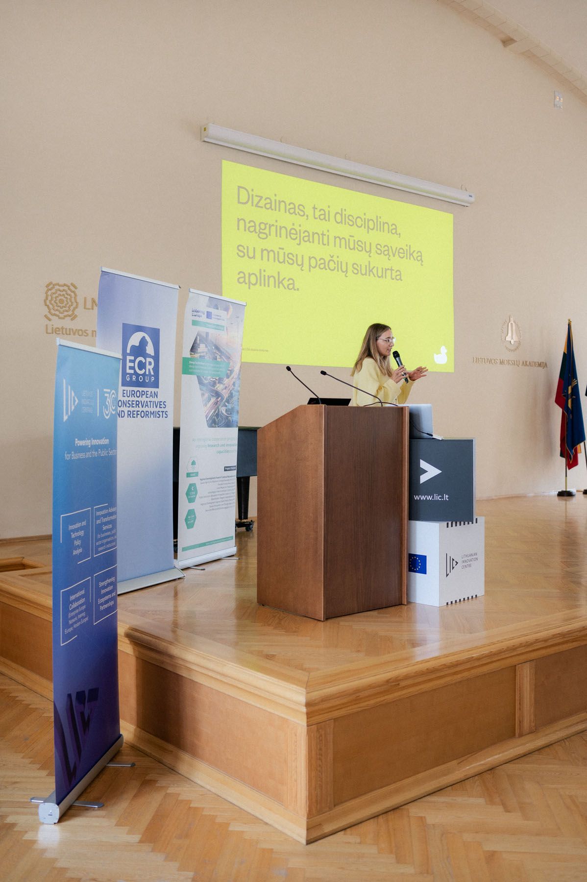 Woman speaks at a wooden podium on stage with a Lithuanian text slide projected behind her and event banners nearby.