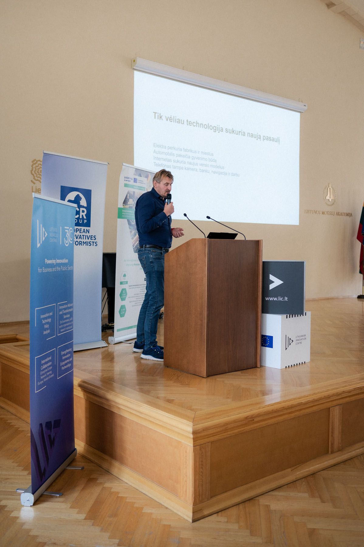 Man holding a microphone presents beside a wooden podium with a projected slide and event banners.