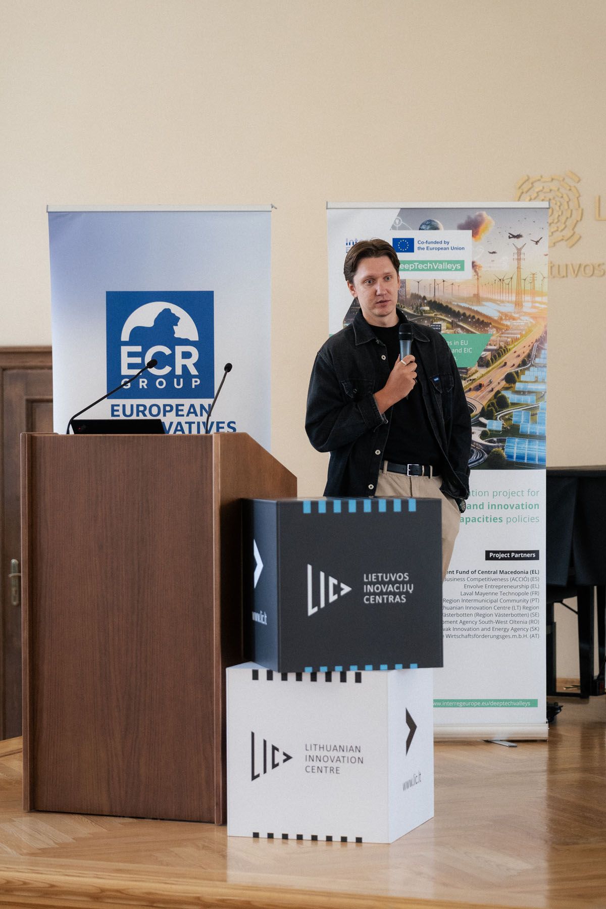 Speaker with a microphone stands beside a wooden podium and Lithuanian Innovation Centre cubes at an ECR Group conference.