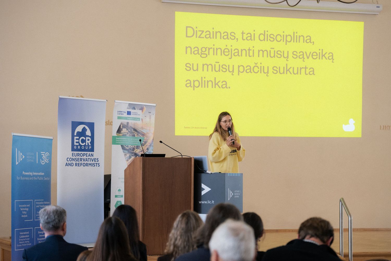 Speaker in a yellow sweater presents on stage with a Lithuanian-language slide about design while the audience watches.