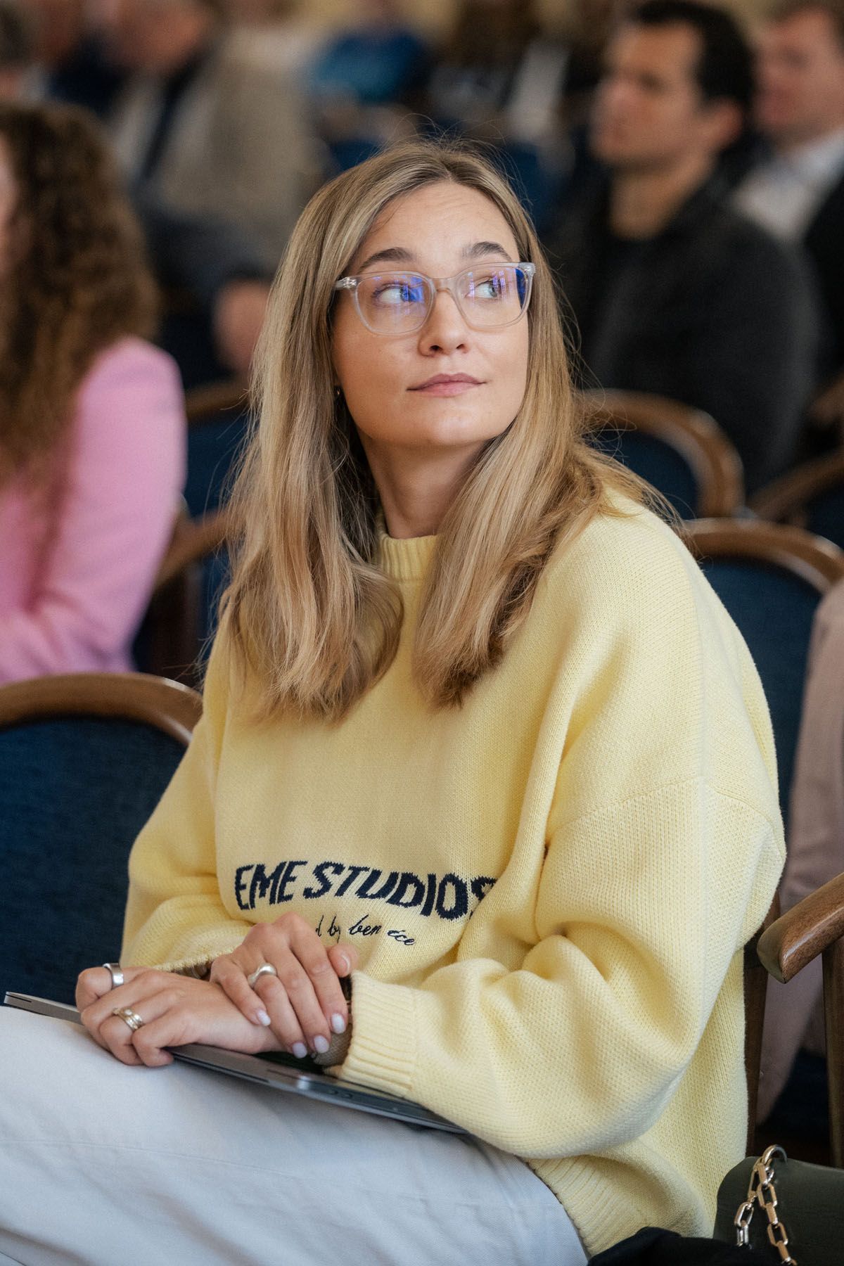 A woman with glasses and a yellow sweater listens thoughtfully while seated among attendees at a conference event.