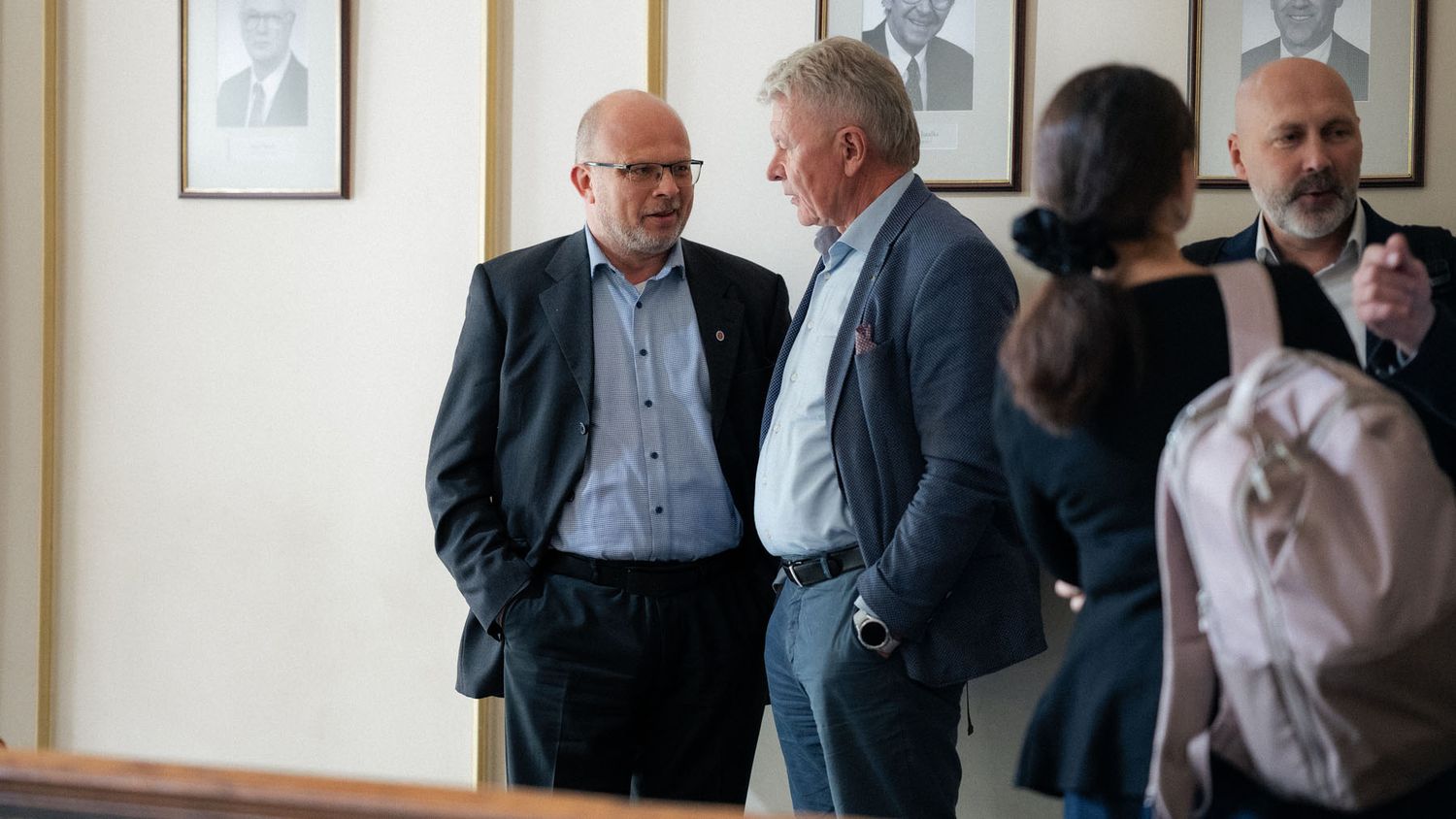 Men in blazers talk in a hallway with framed portraits during a conference networking break.