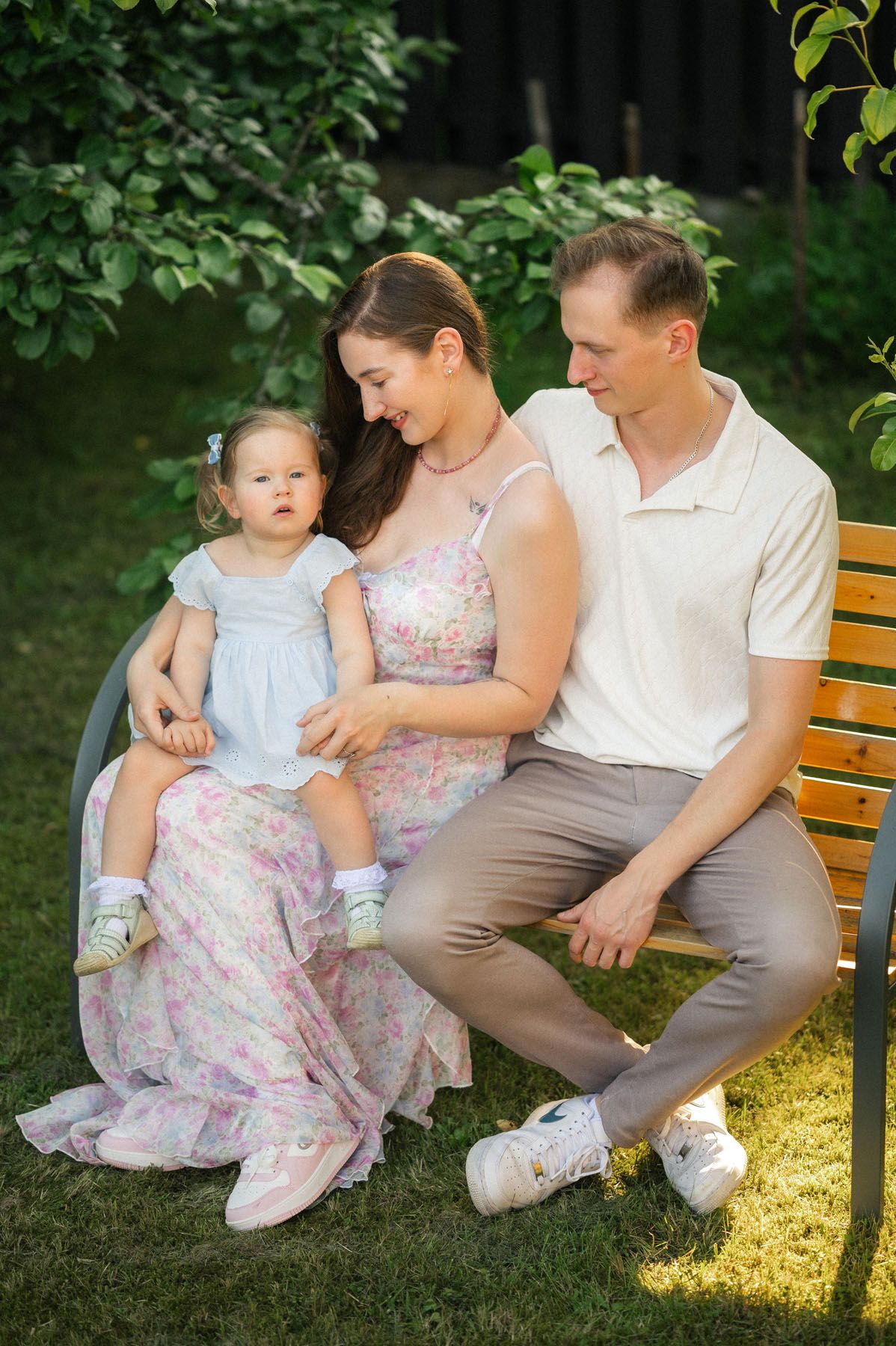 Parents sitting on outdoor bench with toddler daughter, garden family portrait
