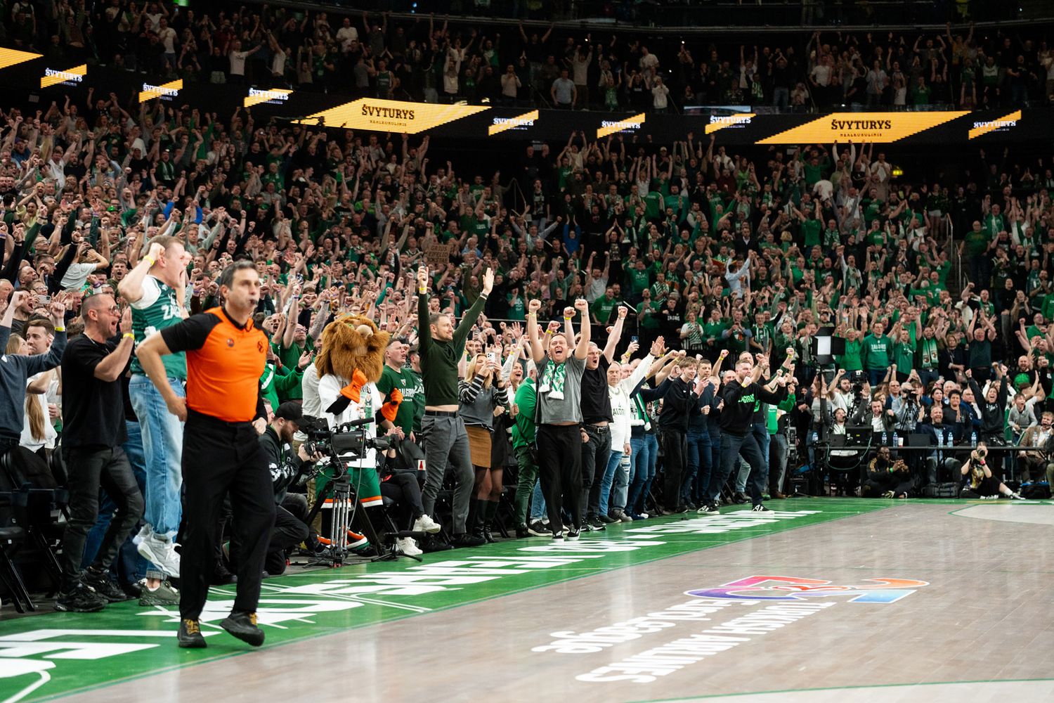 A referee in an orange shirt stands on a basketball court as fans in green cheer and raise their arms in a packed arena.