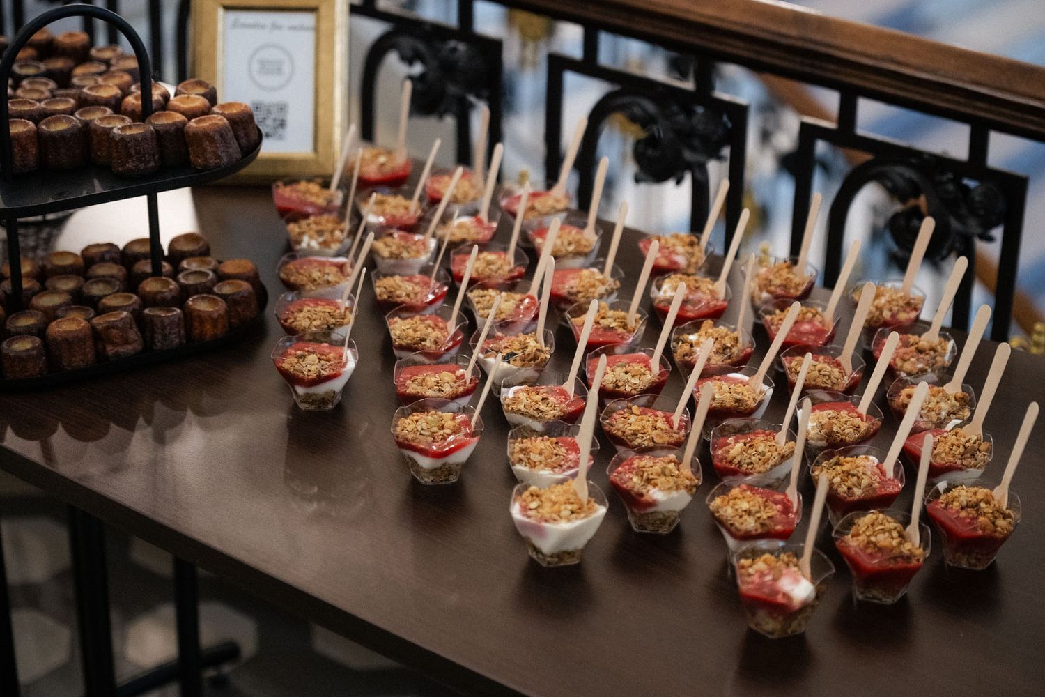 Rows of small glass cups filled with bite-sized appetizers and wooden picks sit on a dark table beside a tray of chocolate desserts.
