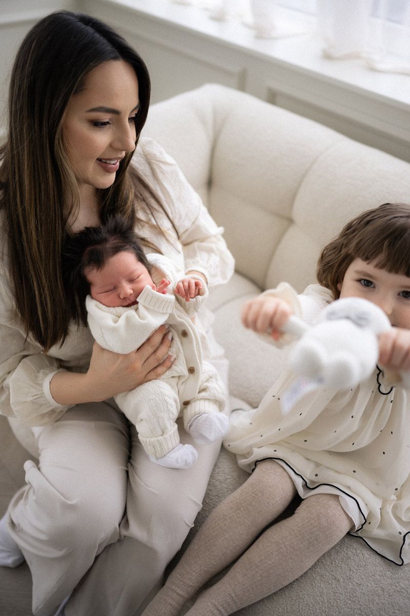 Mother holding newborn baby while older daughter stands beside her, studio family photoshoot