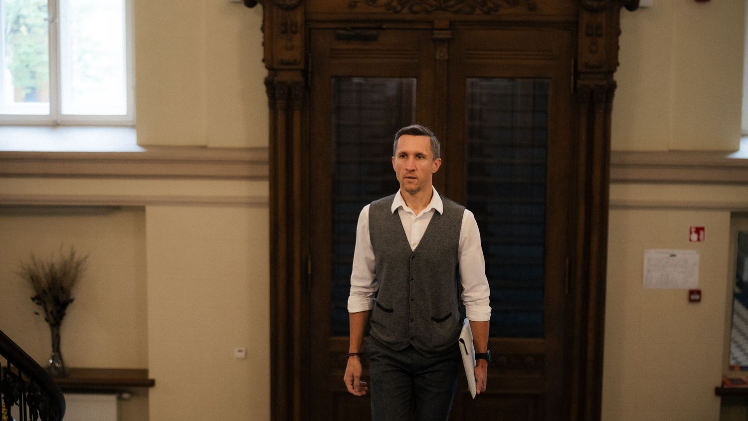 A man in a gray vest and white shirt stands in front of ornate wooden doors in a historic building.