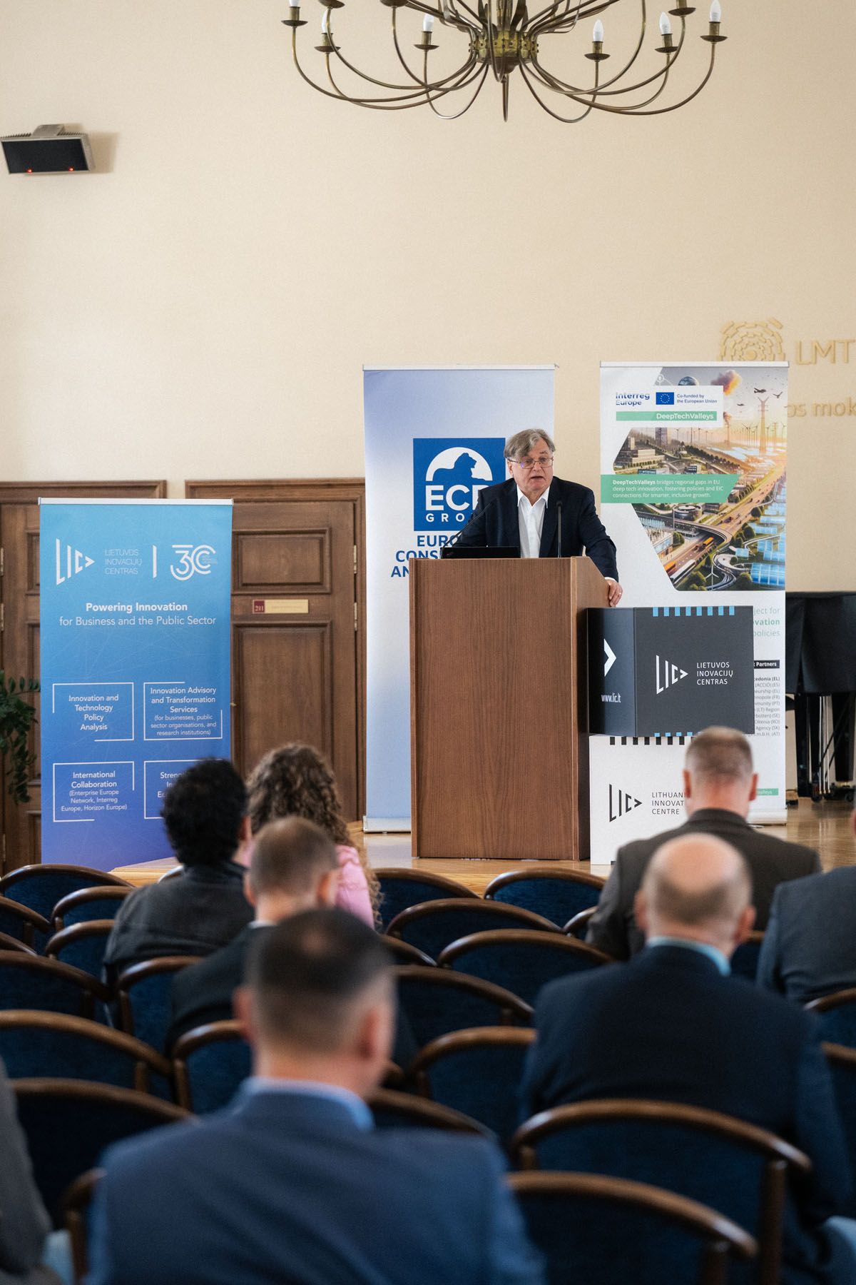 Speaker addresses seated attendees from a wooden podium under a chandelier, flanked by ECR Group and innovation banners.