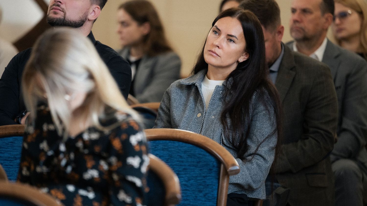 Woman in a gray denim jacket listens attentively among seated conference attendees in a formal hall.