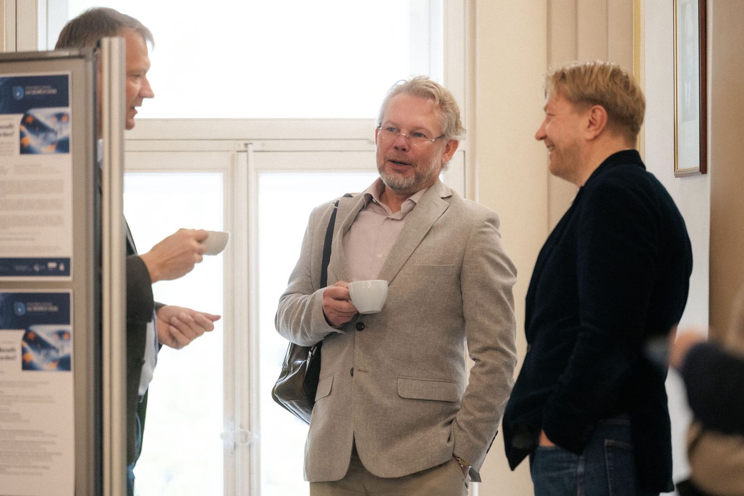 Two men and a woman chat with coffee cups near a bright window and an informational poster stand at a networking event.