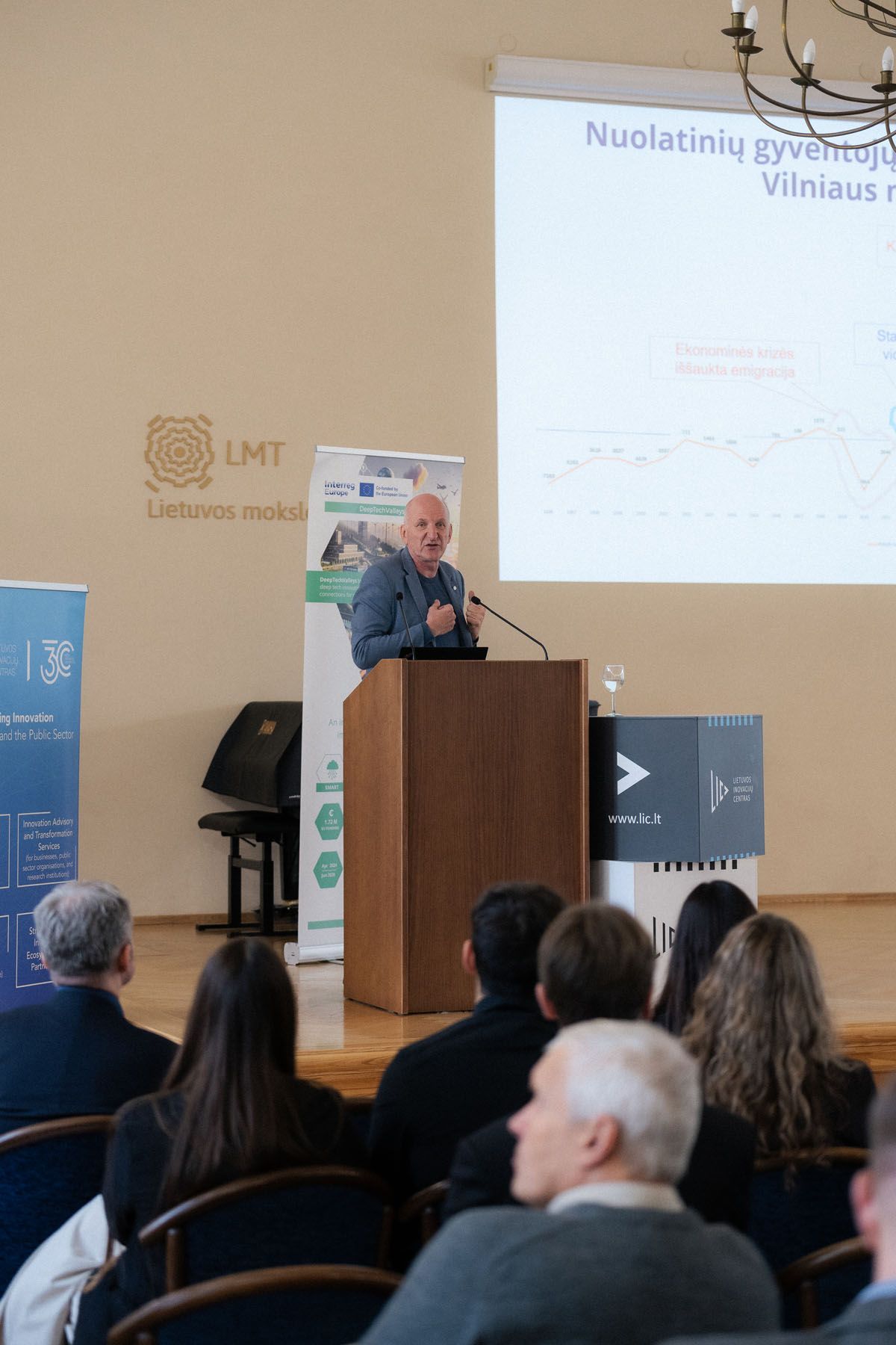 A speaker presents at a podium to a seated audience at a Lithuanian conference with a projected slide behind him.