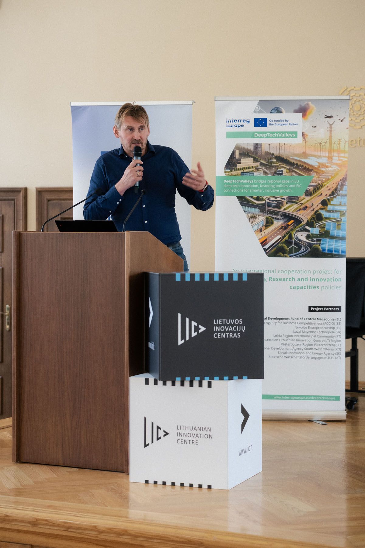 Speaker in a blue shirt holds a microphone at a wooden podium beside Lithuanian Innovation Centre boxes and project banners.