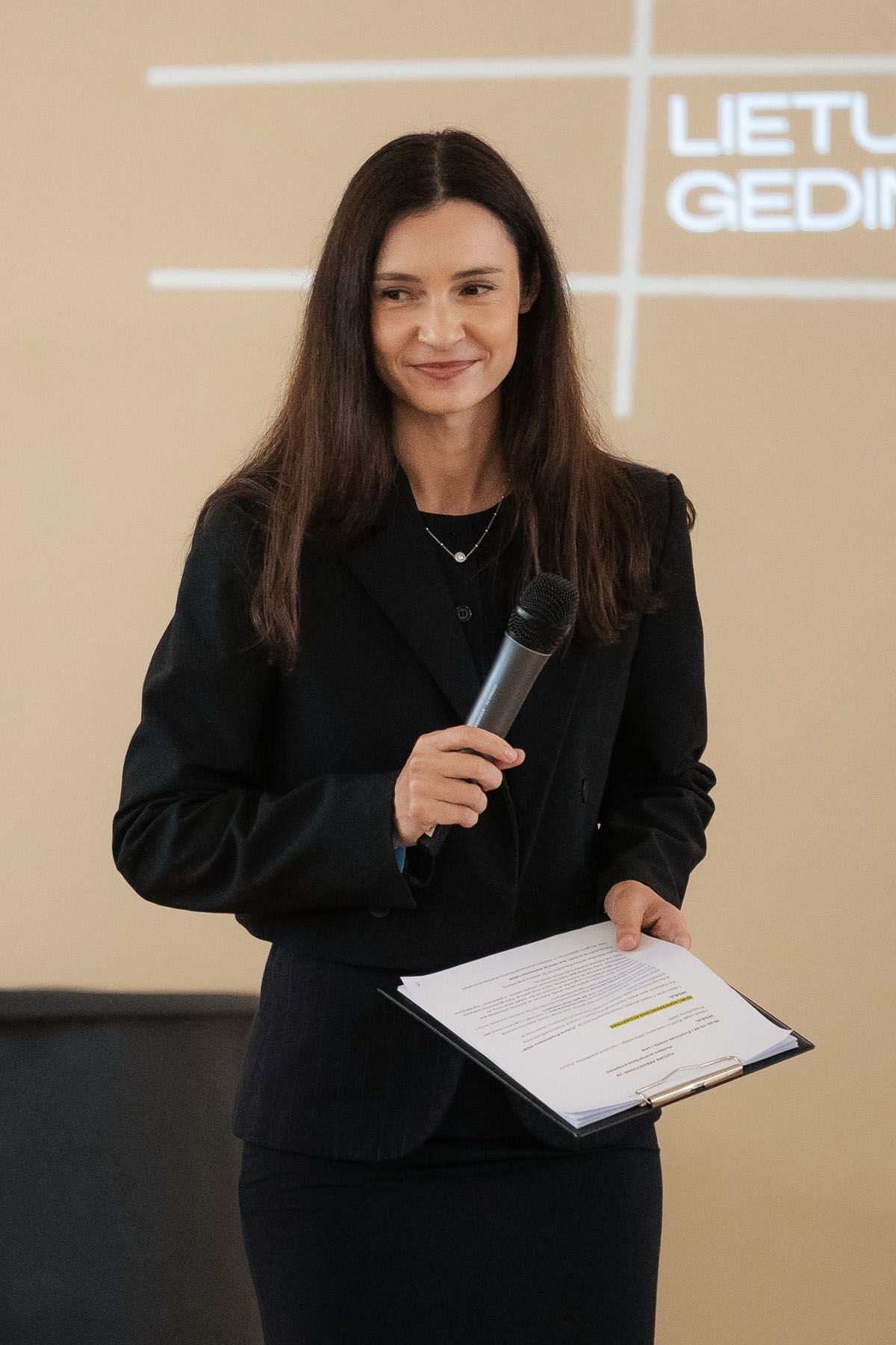 A smiling woman with long dark hair holds a microphone and a clipboard while hosting a conference presentation.