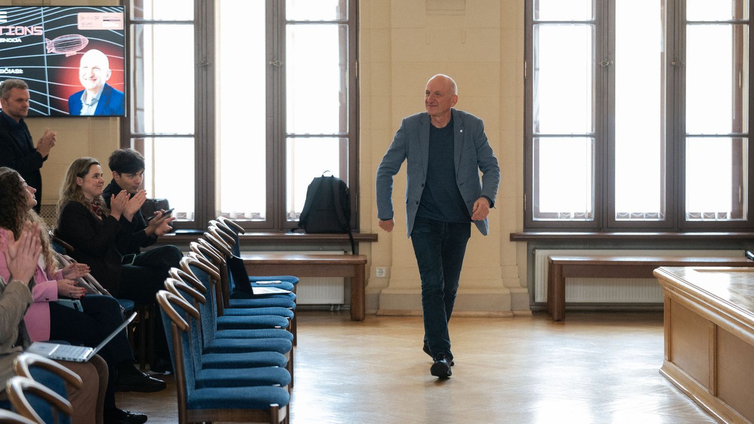 A bald man in a blue blazer walks across a hall past audience seating, with a presentation screen visible at left.