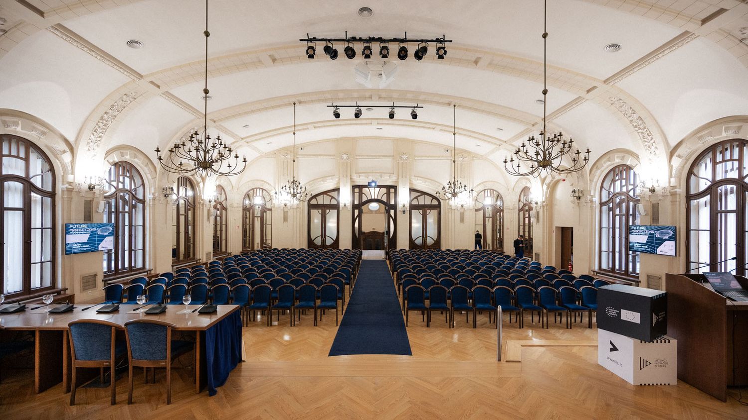 Elegant conference hall with a blue carpeted aisle, rows of seats, crystal chandeliers and tall arched windows.