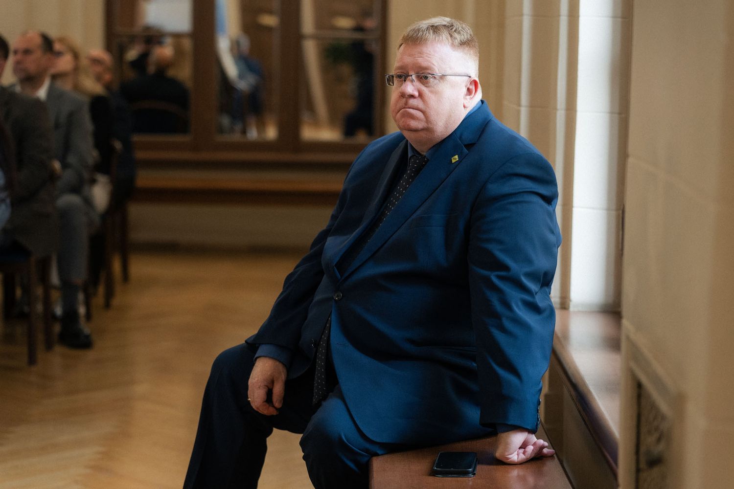 Man in a blue suit and glasses sits on a wooden bench in a bright hall with conference attendees behind him.