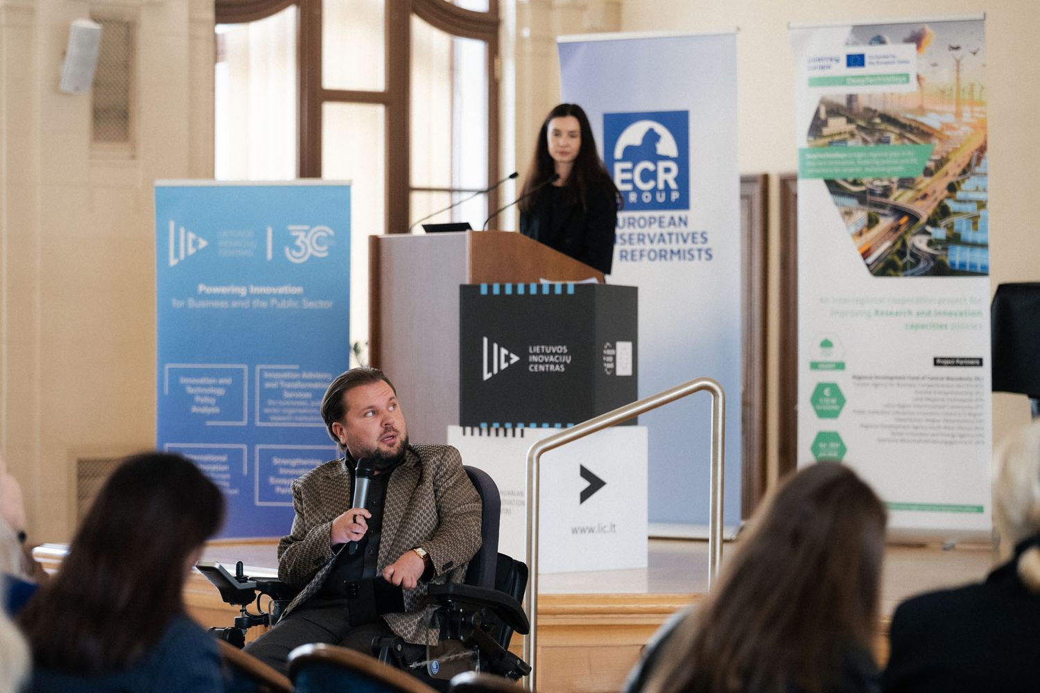 A man in a wheelchair holds a microphone in the foreground while a woman stands at a podium near ECR and Interreg banners.