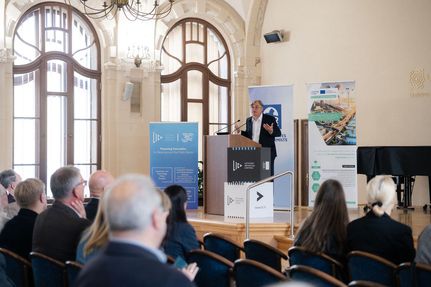Attendees in a grand hall with arched windows listen to a speaker at a podium beside event banners at a conference.