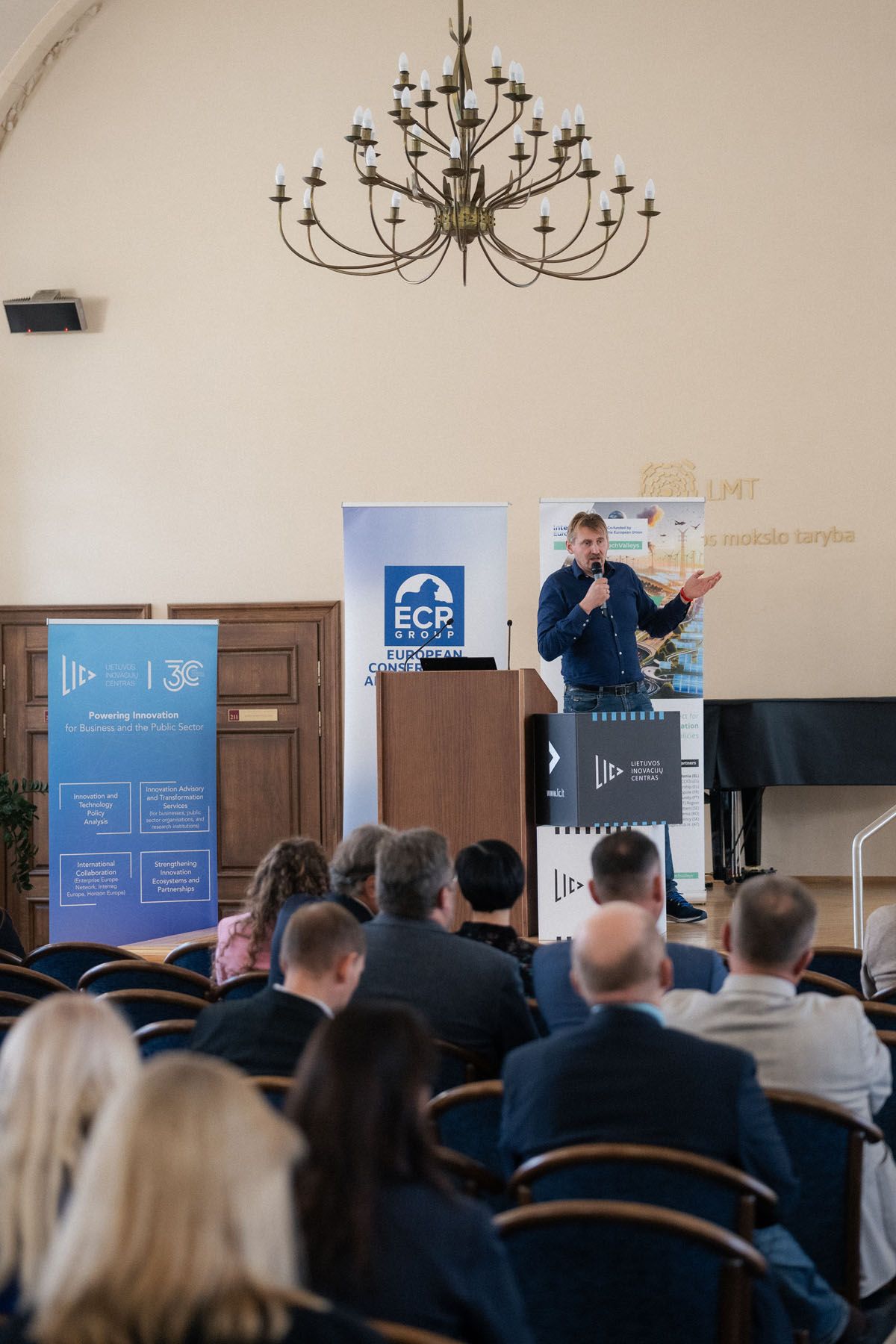 An audience watches a speaker with a microphone at a podium in a hall with a chandelier and Interreg Europe banners.