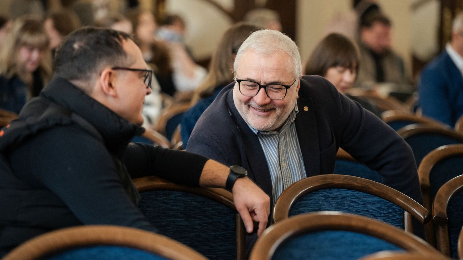 A man with glasses and gray hair smiles while chatting with another man in a seated audience at a conference hall.