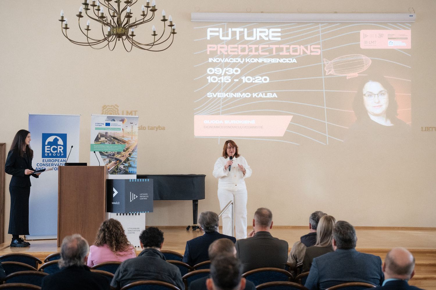Audience seated in a chandelier-lit hall watches a woman in white present Future Predictions as another stands near the podium.