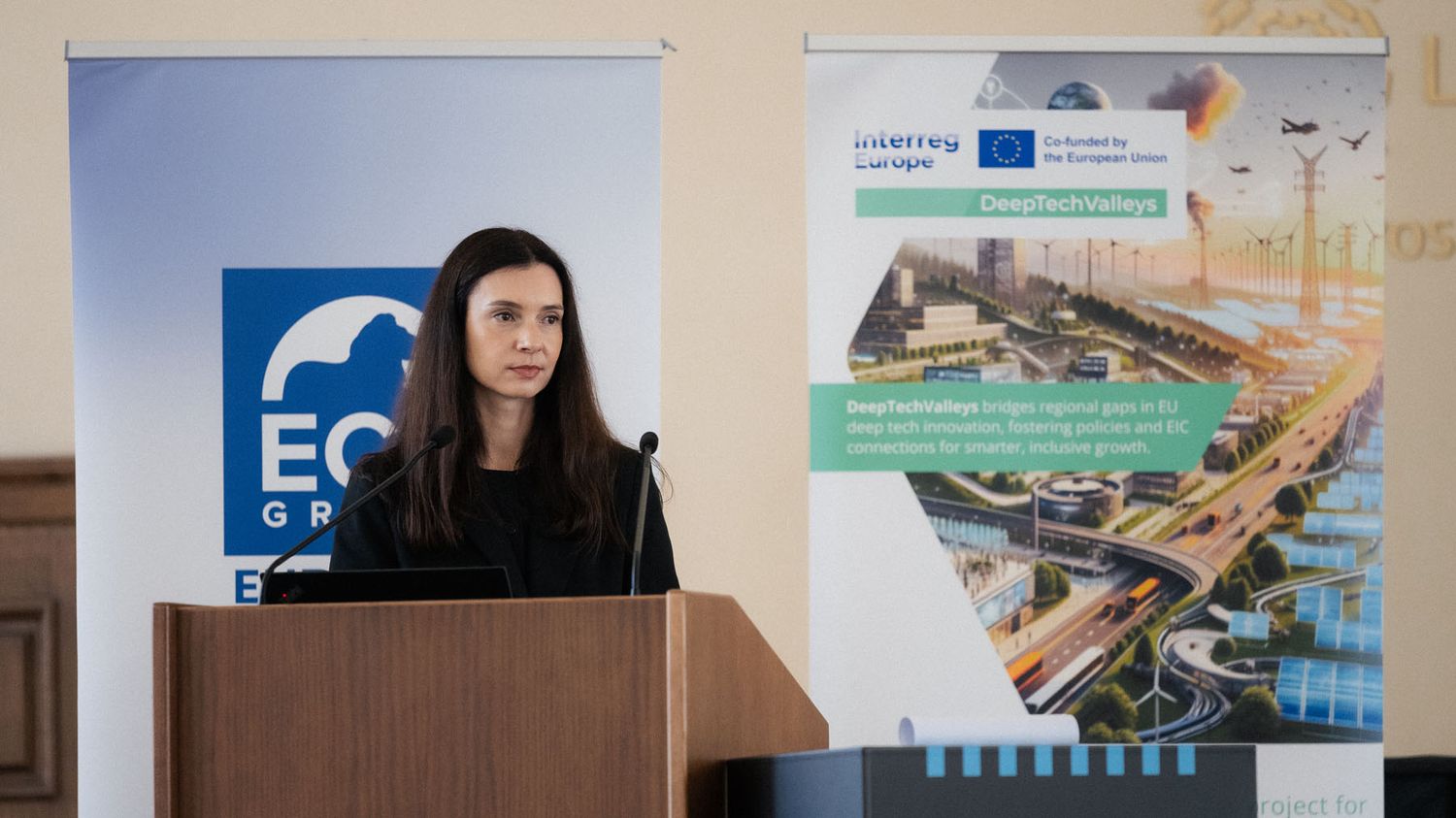 A dark-haired woman stands at a wooden podium with microphones between an ECR banner and a DeepTechVillage Interreg Europe banner.