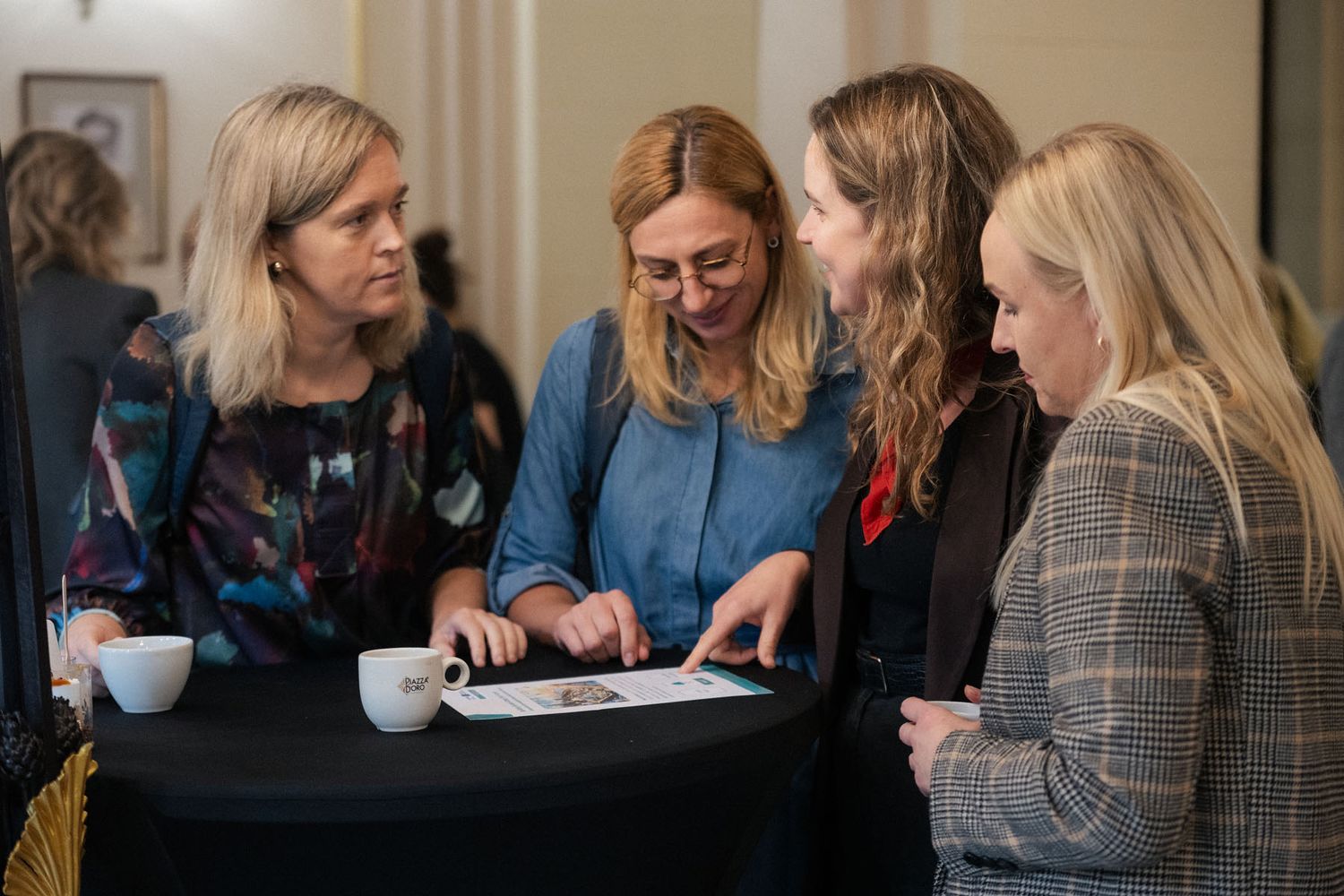 Four women gather around a black cocktail table, reviewing a document and chatting during a conference networking break.