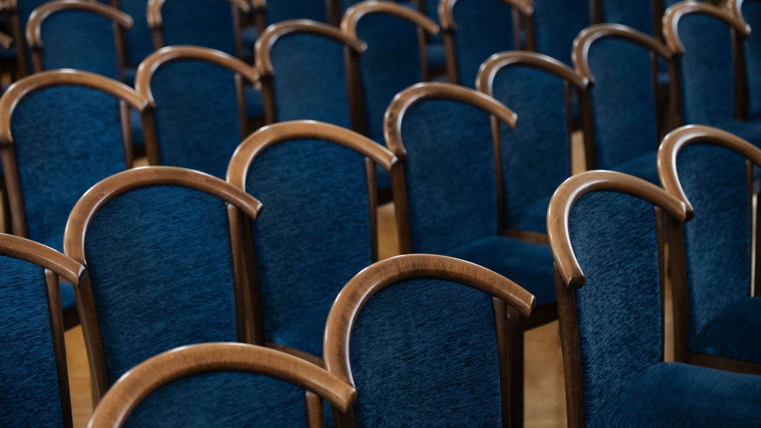 Rows of blue upholstered auditorium seats with curved wooden armrests sit empty in a theater hall.