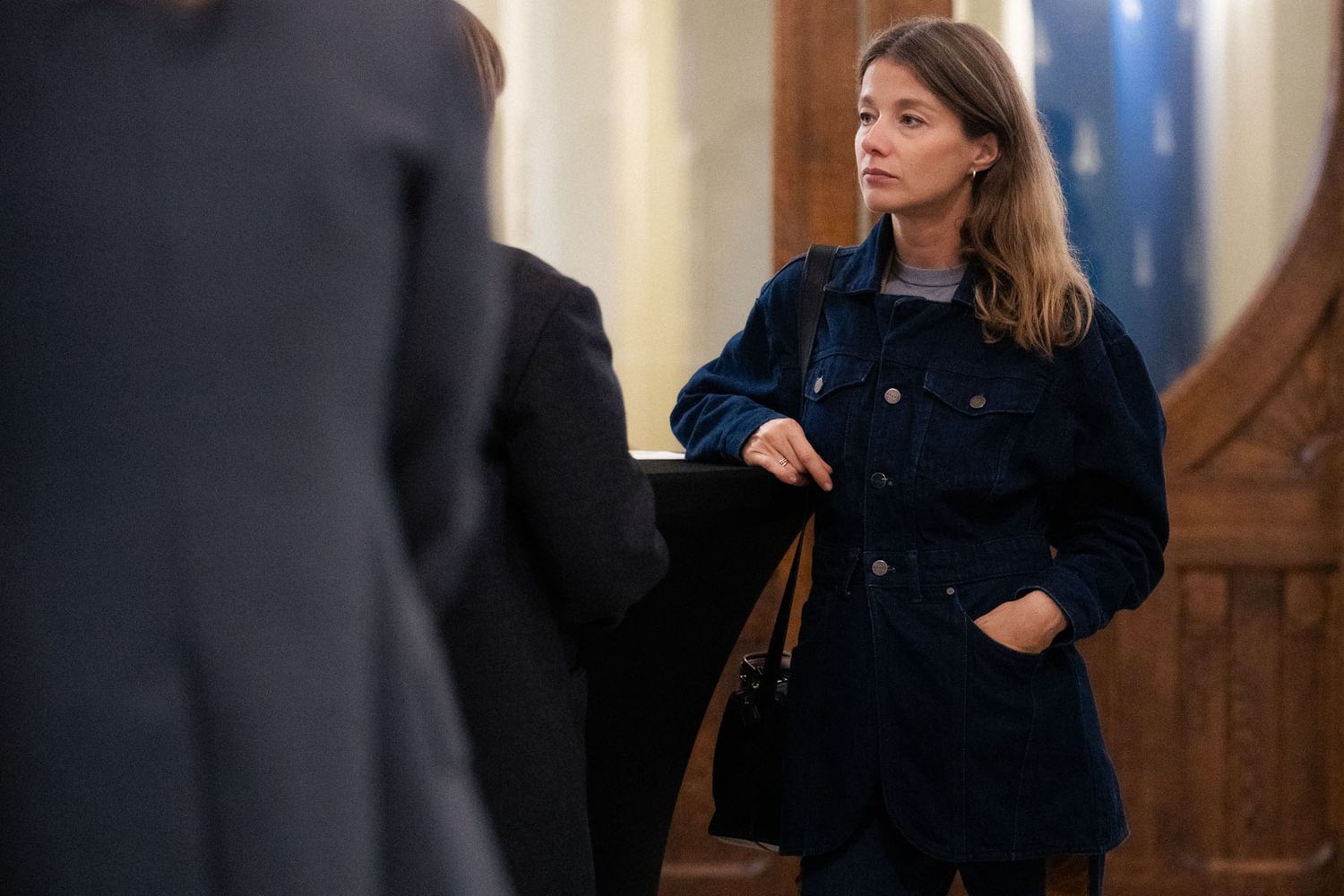A woman in a dark denim jacket stands thoughtfully with a shoulder bag in a wood-paneled room, surrounded by other attendees.