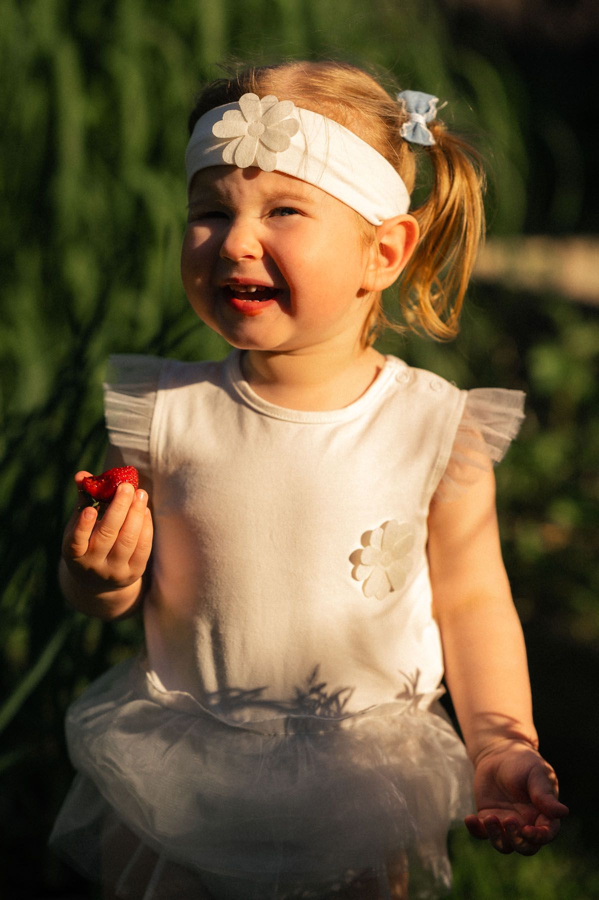 Toddler laughing outdoors in white dress with floral headband, golden light