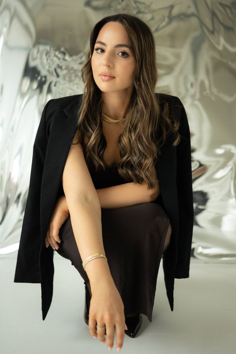 Woman with long wavy hair in a black blazer and gold jewelry crouches in front of a shimmering silver backdrop.
