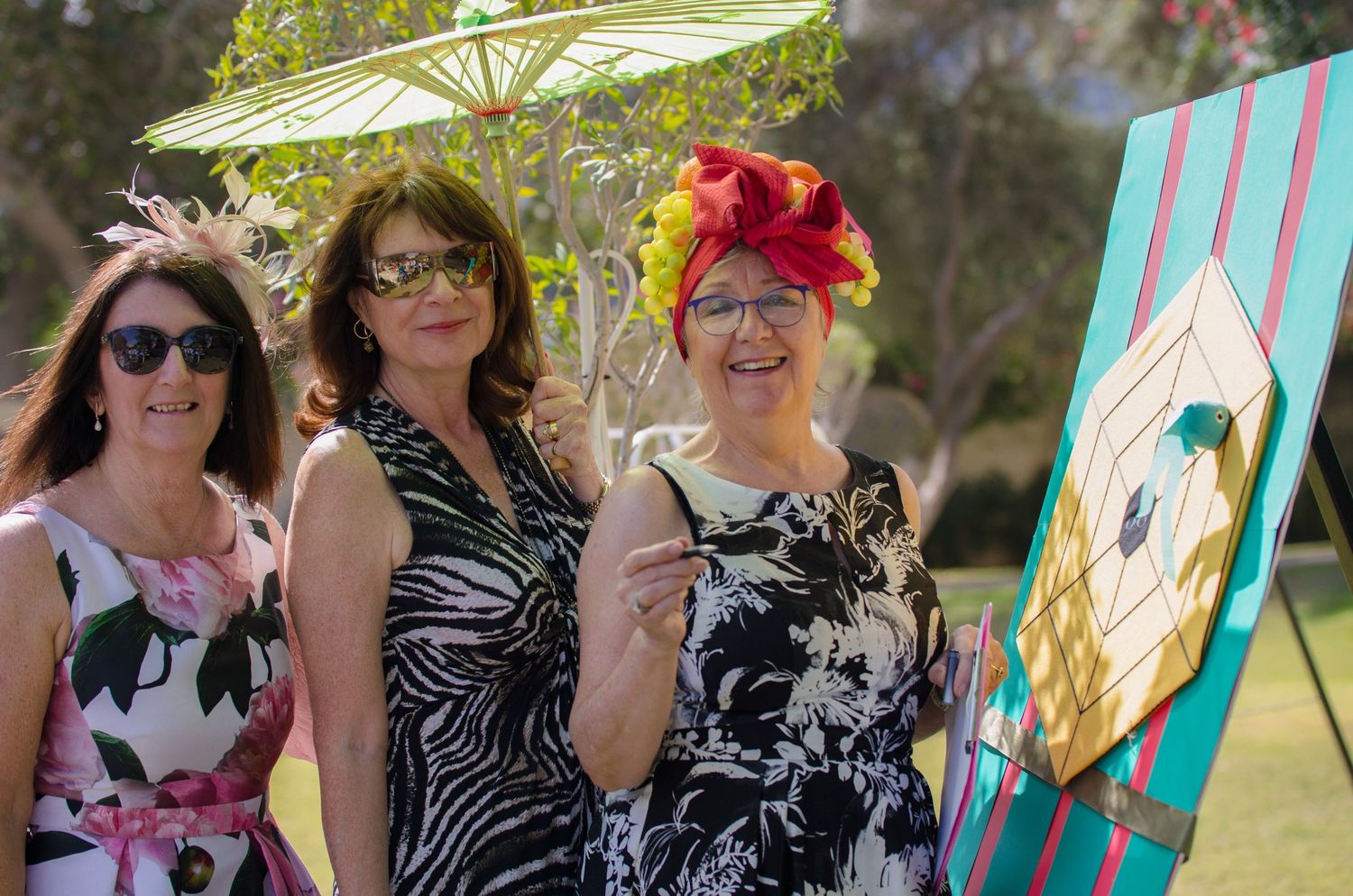 Three smiling women in elegant floral dresses and fascinators pose outdoors beside a colorful painted board and a decorative parasol.