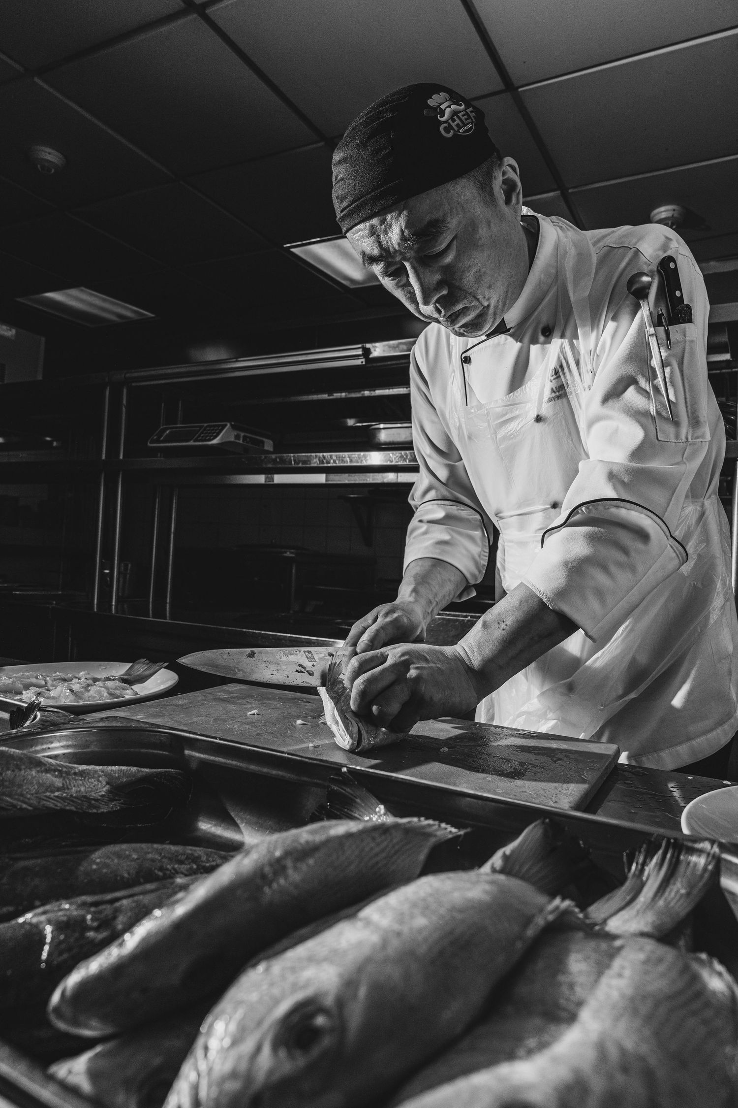 Black and white photo of a chef in white uniform filleting fresh fish in a professional kitchen.