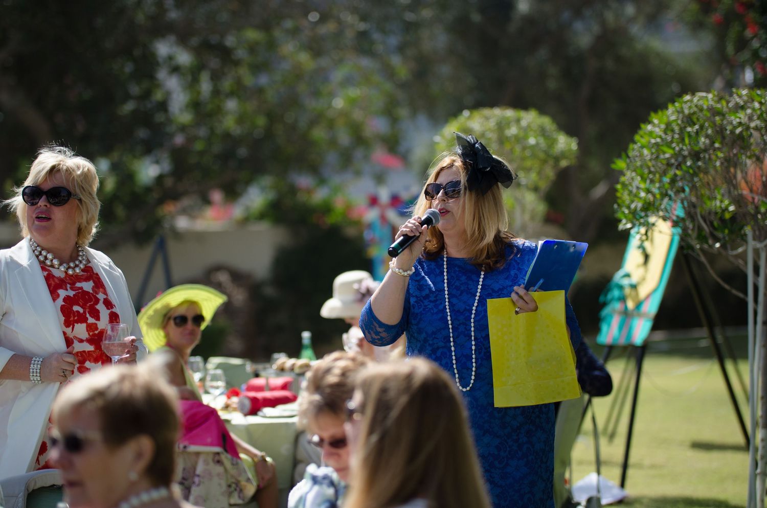 Woman in a blue dress and black fascinator speaks into a microphone holding a yellow bag at an outdoor garden party with seated guests.