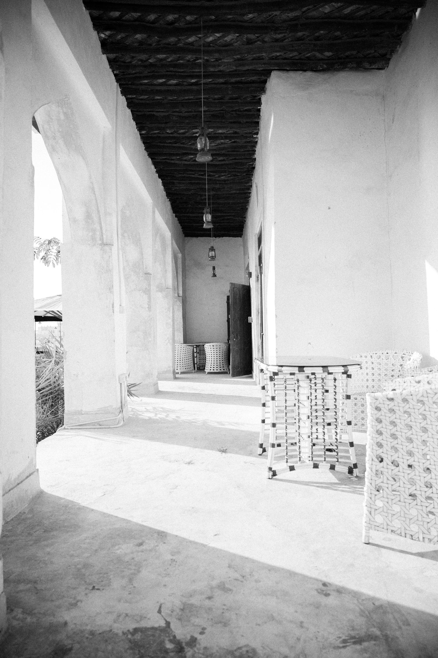 Black and white photo of a sunlit corridor with white walls, dark ceiling, hanging lamps, and ornate furniture.