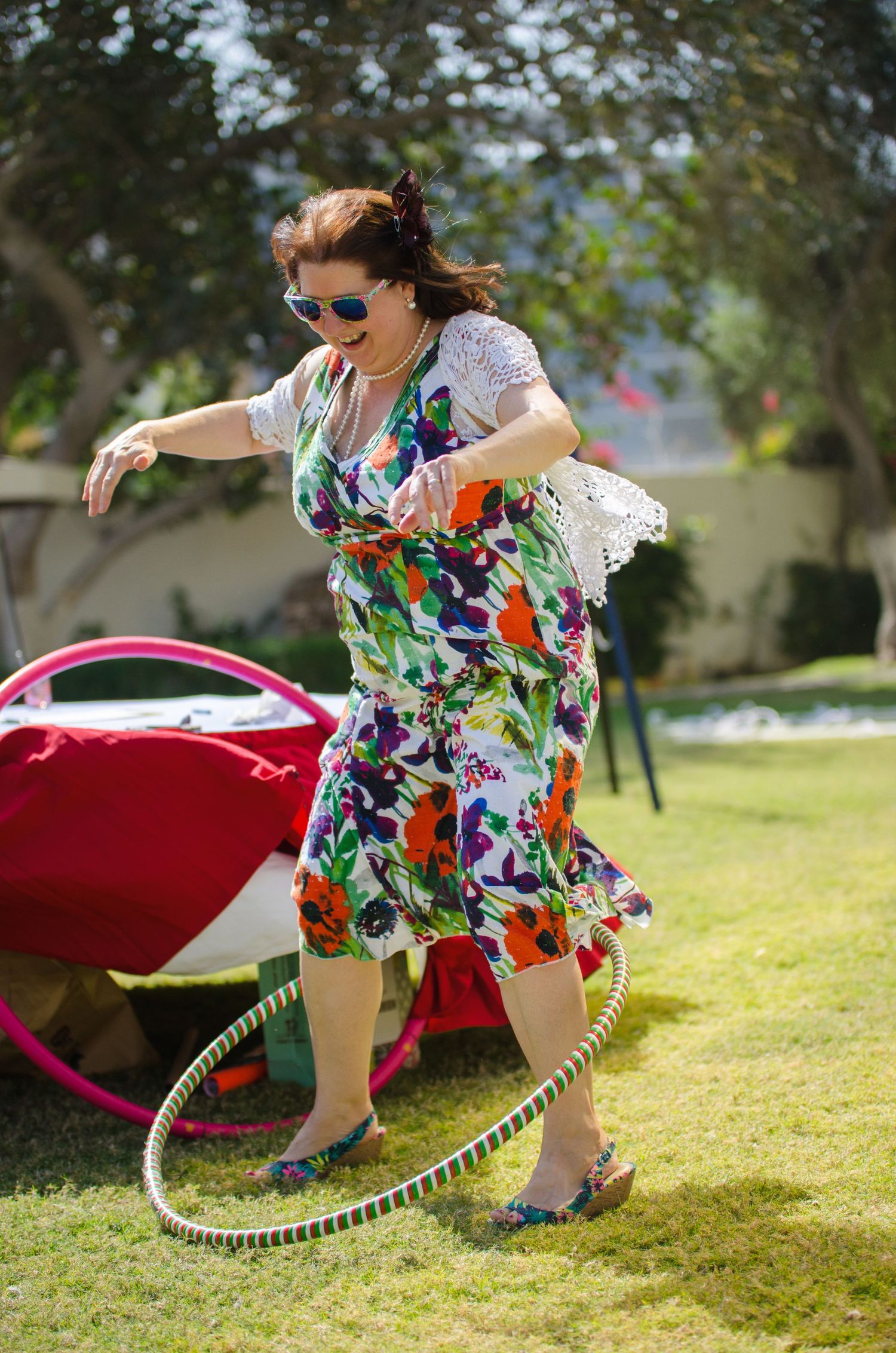 Woman in sunglasses and a colorful floral dress spins a hula hoop on a sunny green lawn at an outdoor party.
