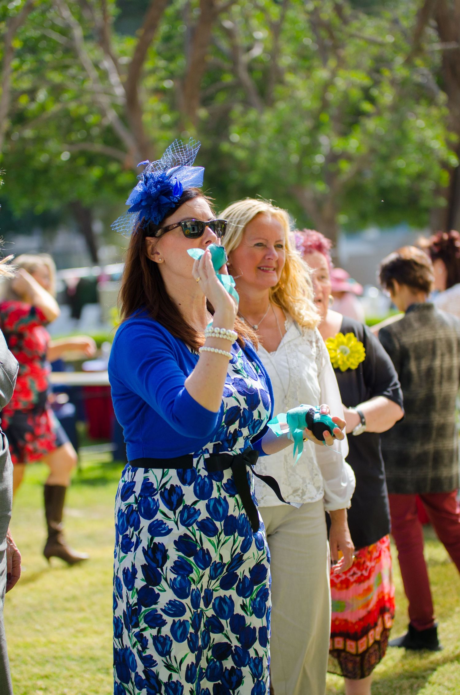 Woman in a blue floral dress, sunglasses and blue fascinator holds a phone at an outdoor event with elegantly dressed guests on the lawn.