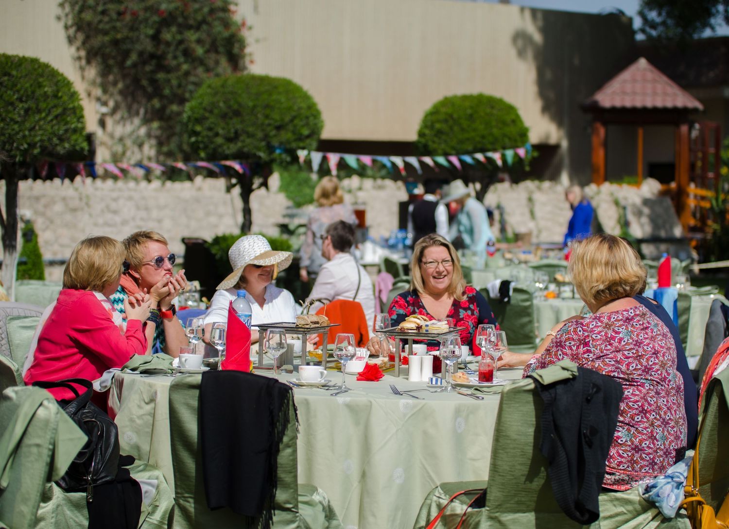 Group of women sit around an outdoor table with food and drinks, chatting during a sunny garden party with bunting and trimmed trees.
