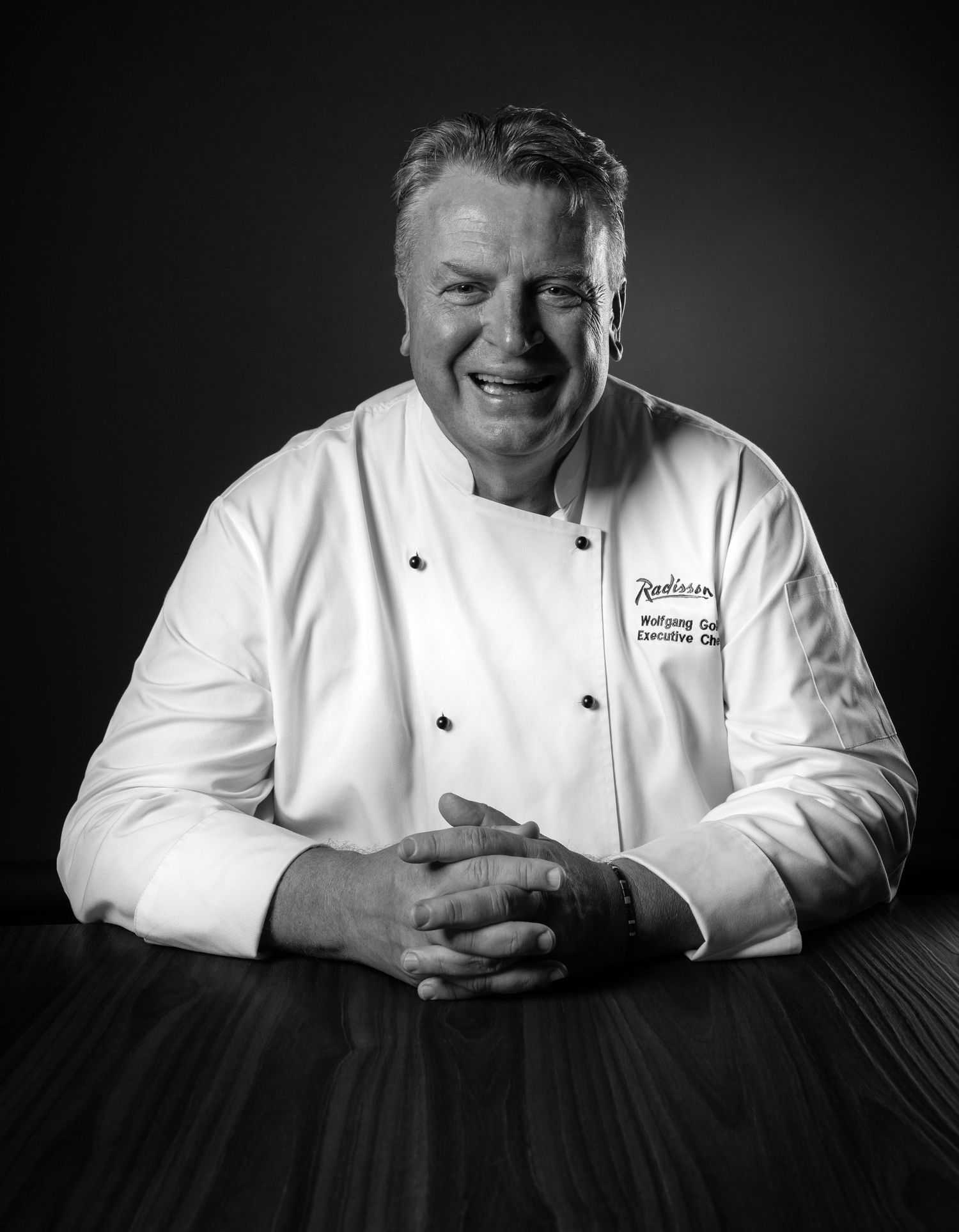 Black and white portrait of a smiling executive chef in white uniform with Radisson logo, seated at a wooden table.