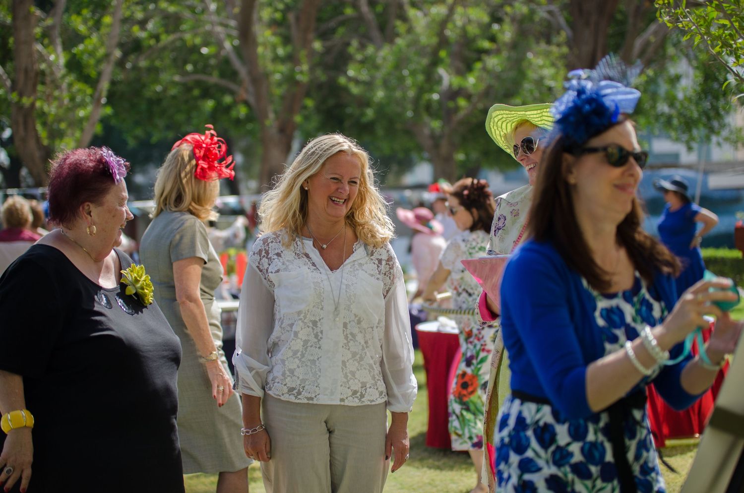 Women in colorful fascinators and spring dresses mingle at an outdoor garden party beneath tall shady trees.