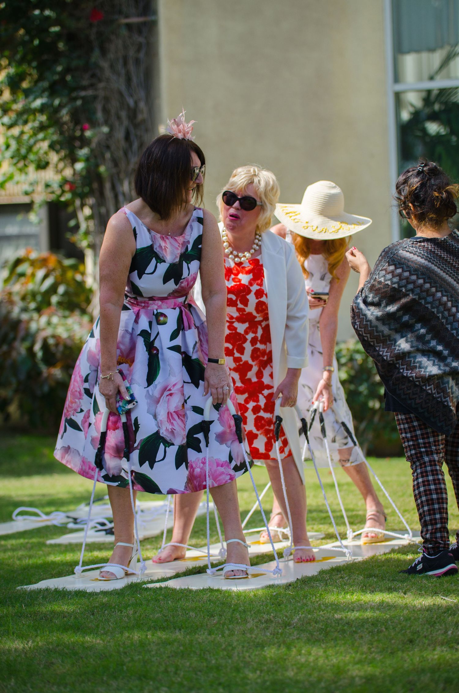 Women in floral dresses and sun hats balance on small mats in a garden while playing a lively outdoor party game on the grass.