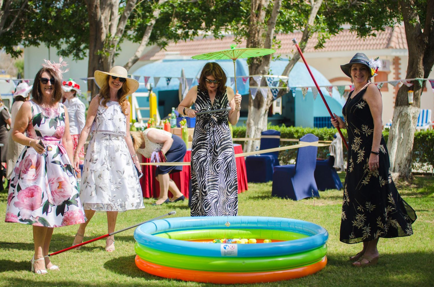 Women in summer dresses and sun hats stand around a colorful inflatable pool at an outdoor garden party under shady trees.