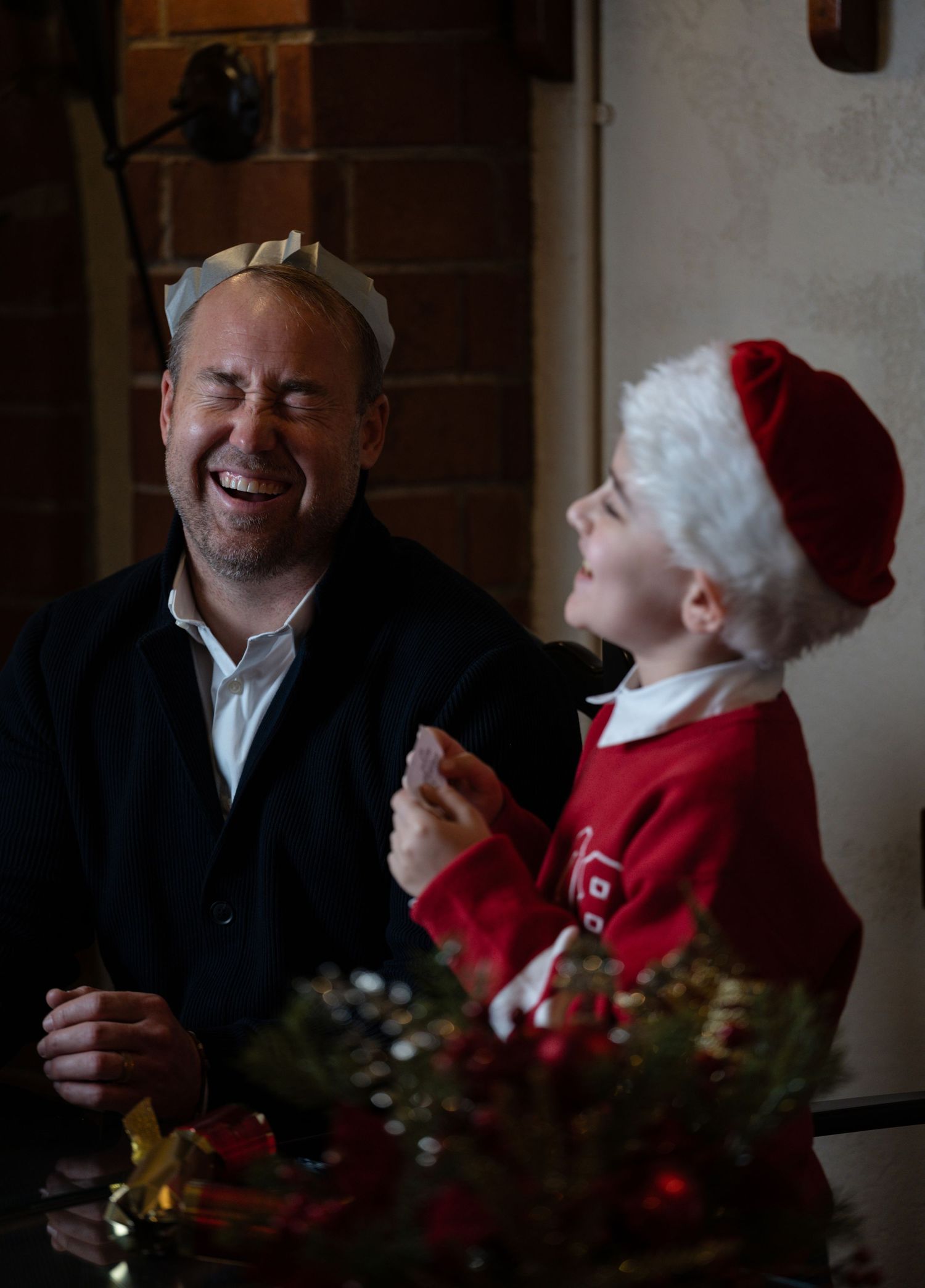 A man and child laugh together at a Christmas table, both wearing festive hats, sharing a joyful holiday moment.