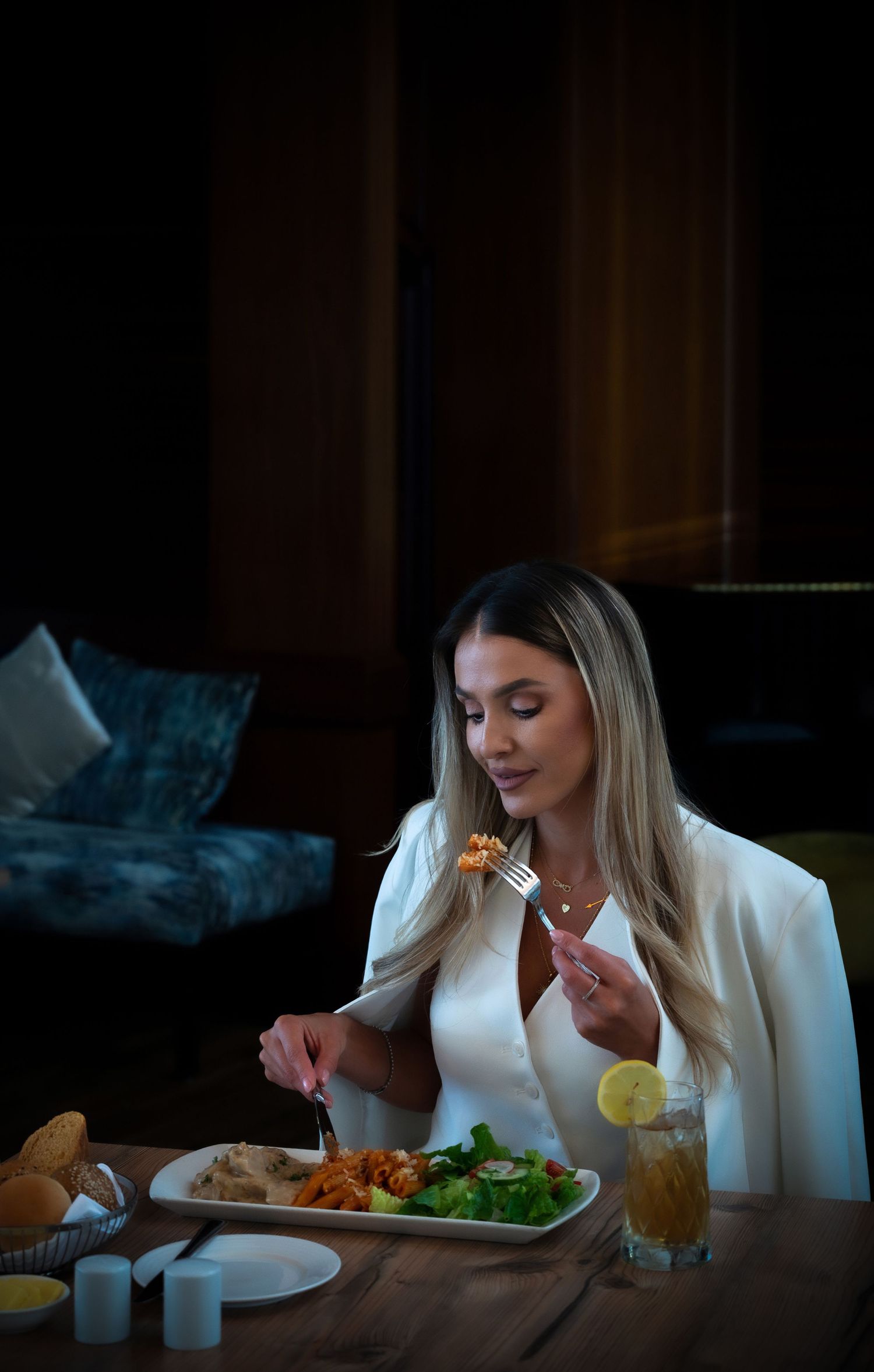 Elegant woman in white blazer dining at upscale restaurant, enjoying a meal with salad and iced tea.