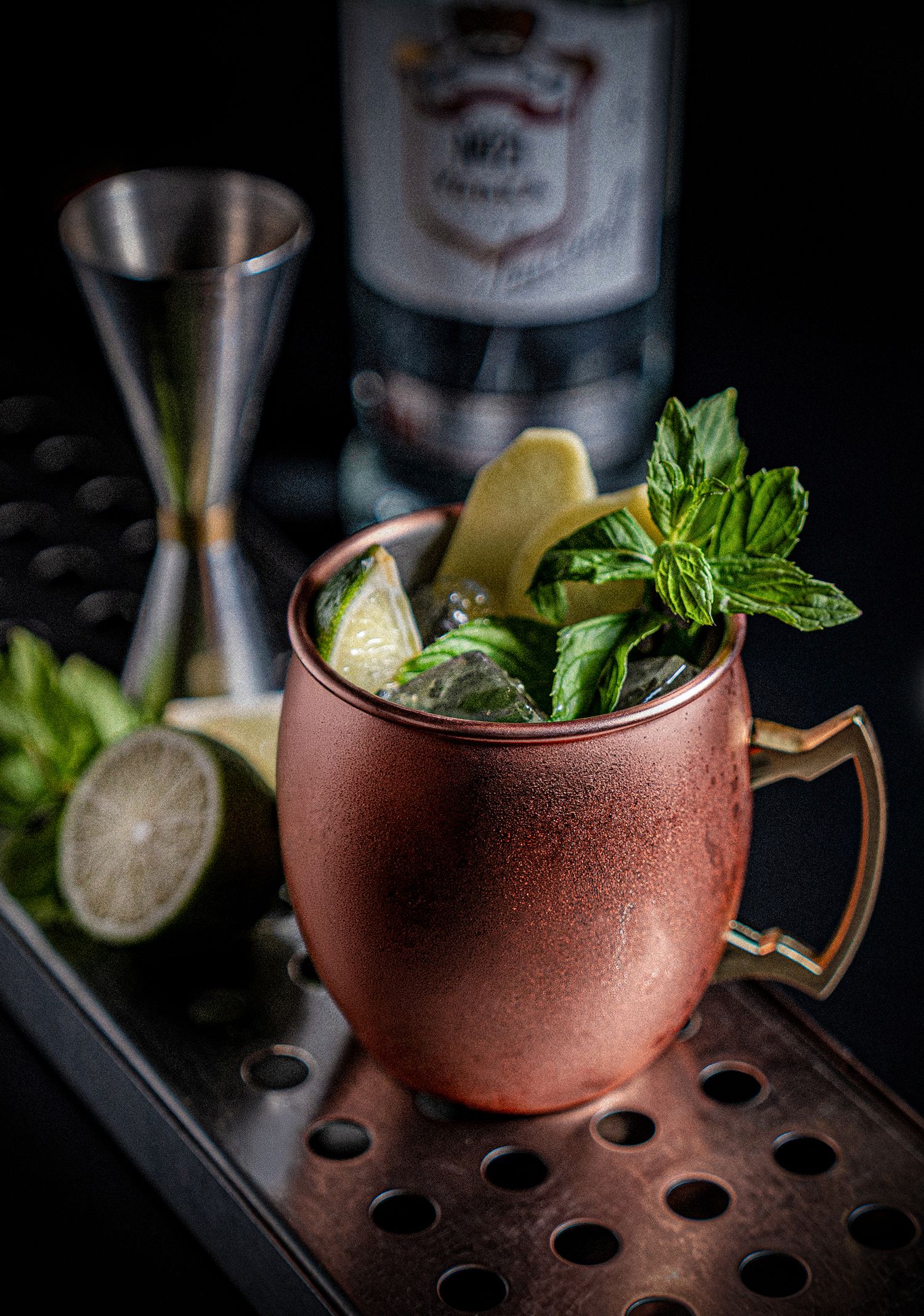 Moscow Mule cocktail in copper mug with fresh mint, lime wedges, ice, jigger and bottle on dark background.