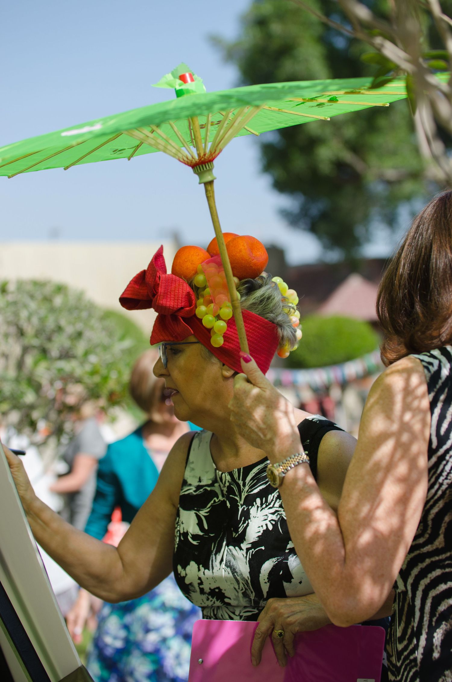 Woman in a red bow and fruit-topped hat holds a green parasol with a toy parrot at a sunny outdoor garden party.