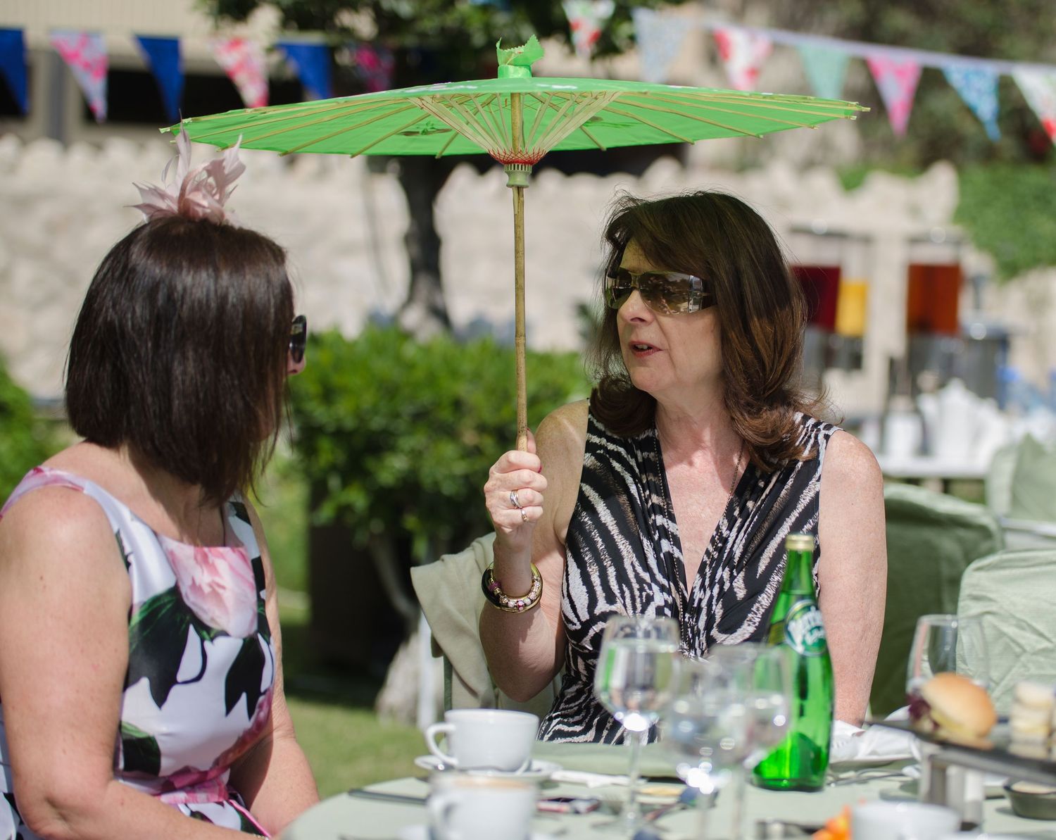 Two women sit at an outdoor table with tea cups and glasses while one holds a small green parasol, with festive bunting in the background.